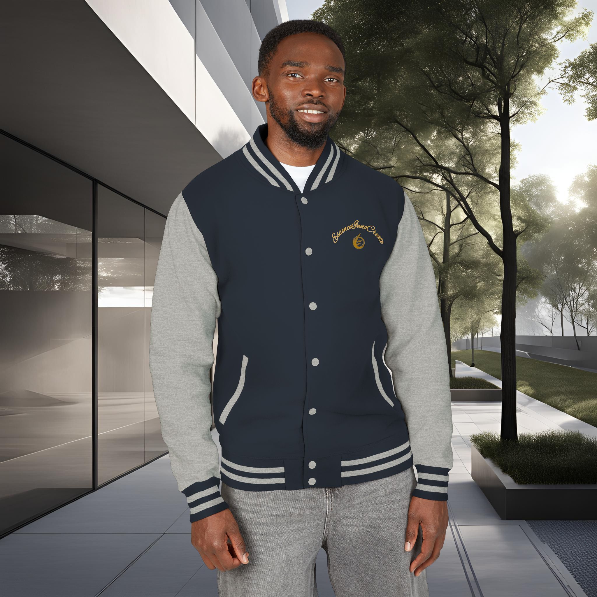 13396497876151231691_2048_customjpeg - Essence Innovation Man wearing a navy and gray varsity jacket stands outdoors in front of a modern glass building and trees, smiling slightly at the camera.