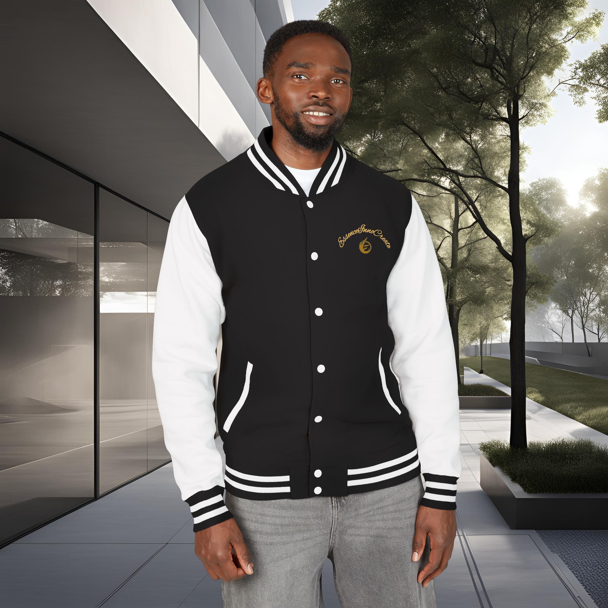 15875421397053886541_2048_customjpeg - Essence Innovation Man wearing a black and white varsity jacket stands outdoors in front of a modern glass building with trees in the background, smiling at the camera.