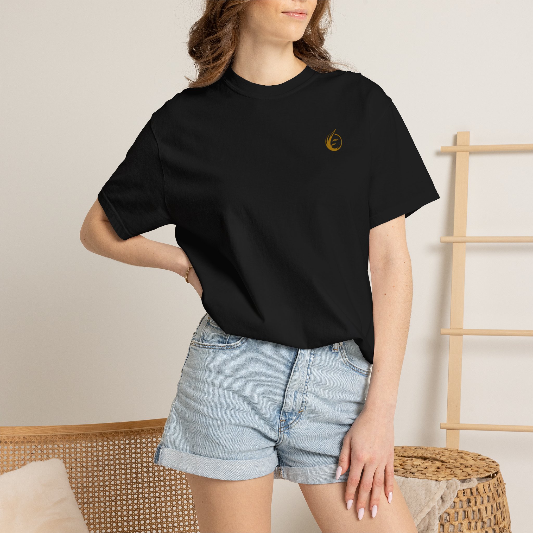 5598203533526772150_2048jpeg - Essence Innovation Woman wearing a black T-shirt with a small gold logo on the chest, paired with light denim shorts, standing near a wicker bench and wooden ladder.